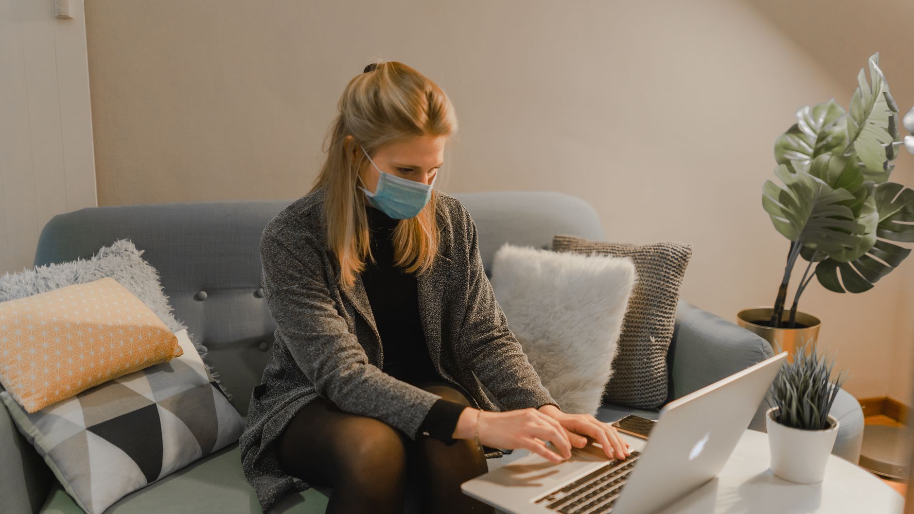 Une femme portant un masque, assise sur un canapé bleu-gris entouré de coussins, tape sur un ordinateur portable.